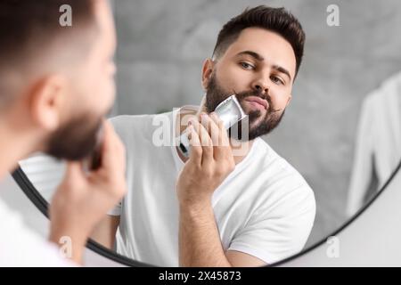 Handsome young man trimming beard near mirror in bathroom Stock Photo