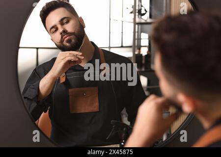Handsome young man trimming beard with scissors near mirror indoors Stock Photo