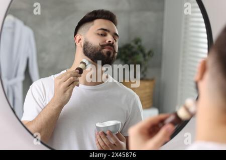 Handsome young man shaving beard near mirror in bathroom Stock Photo