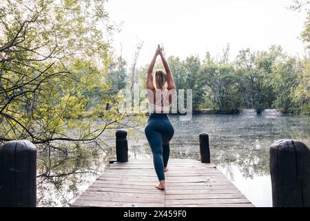 Rear view of a young and athletic woman doing yoga in nature. She is barefoot on a wooden platform near a lake Stock Photo