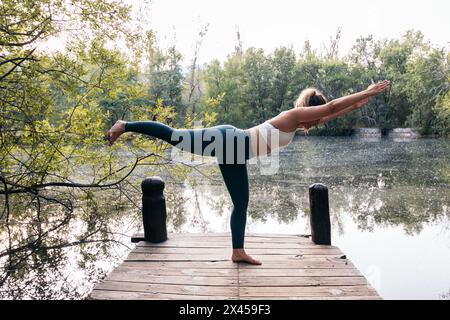 Young woman in sportswear doing yoga in nature. She is barefoot on a wooden platform near a lake Stock Photo