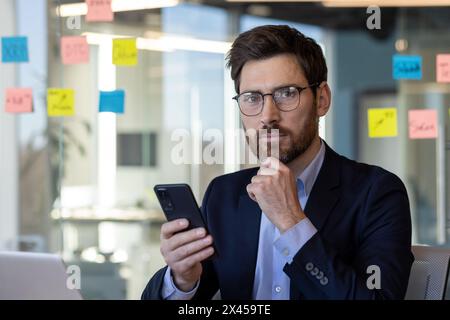 Mature businessman in a modern office setting using a smartphone. He appears thoughtful, wearing glasses and a dark suit, surrounded by colorful sticky notes. Stock Photo