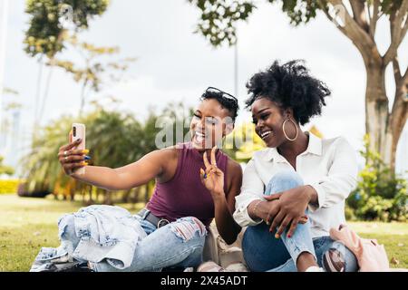 Two black women friends having fun and taking selfies with smartphone sitting on the grass in the park Stock Photo