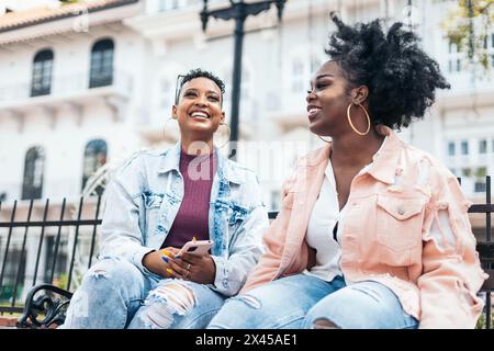 Two black women friends talking sitting on a bench laughing and having fun in the city Stock Photo