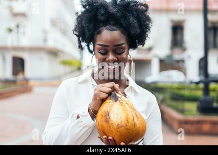 Portrait of an afro-haired black Latin American woman with large earrings holding and drinking a coconut. She is happy and smiling. Stock Photo
