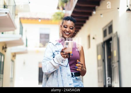 Cool smiling woman with short curly hair and big earrings walking on the city street and using smartphone. Stock Photo
