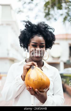 Portrait of an afro-haired black Latin American woman with large earrings holding and drinking a coconut. She is happy and smiling. Stock Photo