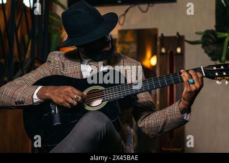 Stylish black man in hat and classic outfit playing guitar indoors Stock Photo