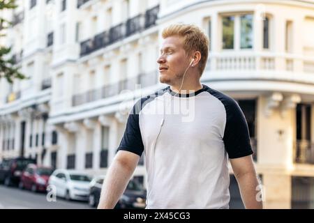 Albino latino man dressed in white clothes walking in the city street listening to music with headphones. His long white lashes shield his eyes from t Stock Photo