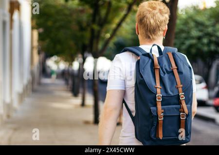 Rear view of a young Latino man who is albino walking down the city street on a summer morning. He wears white clothes and a blue backpack. Stock Photo
