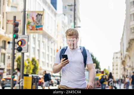 Young albino man in white clothes walking down the street in the city while using smartphone and listening to music with earphones Stock Photo