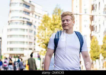 Albino latino man dressed in white clothes and backpack walking in the city street listening to music with headphones. His long white lashes shield hi Stock Photo