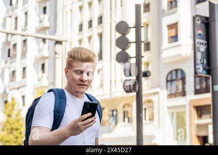 Young albino man in white clothes walking down the street in the city while using smartphone and listening to music with earphones Stock Photo
