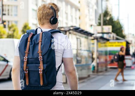 Young Latino man who is albino walking down the city street listening to music with headphones on a summer morning. He wears white clothes and a blue Stock Photo