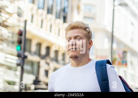 Albino latino man dressed in white clothes and backpack walking in the city street . His long white lashes shield his eyes from the sunlight. Stock Photo