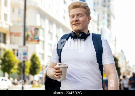 Young Latino man who is albino walking down the city street listening to music with headphones on a summer morning. He wears white clothes and a blue Stock Photo