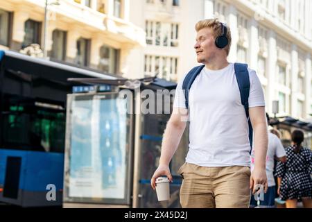 Young Latino man who is albino walking down the city street listening to music with headphones on a summer morning. He wears white clothes and a blue Stock Photo