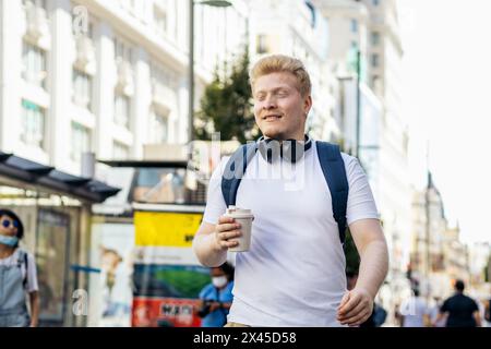 Young Latino man who is albino walking down the city street listening to music with headphones on a summer morning. He wears white clothes and a blue Stock Photo