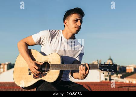 young man sitting playing acoustic guitar on a rooftop Stock Photo