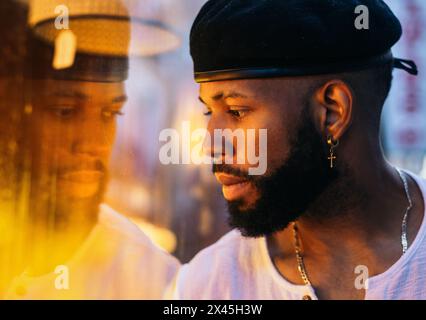 Bearded stylish black man looking with intense gaze through a shop window. You can see his reflection in the glass and there is a warm dim light Stock Photo