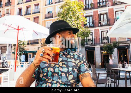 Stylish black man with painted nails and a hat having a drink sitting on a terrace in the city of Madrid on a summer day Stock Photo