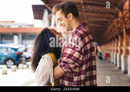 Multiracial young adult couple hugging and kissing after meeting again at the station. She is carrying a blue suitcase from her trip and he waits for Stock Photo