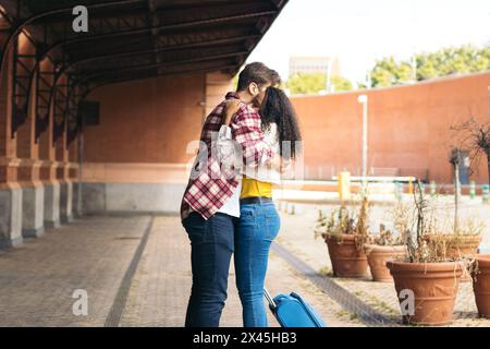 Multiracial young adult couple hugging and kissing after meeting again at the station. She is carrying a blue suitcase from her trip and he waits for Stock Photo