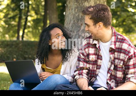 Young couple of multiracial students sitting in the park using a laptop. They work together and have fun Stock Photo