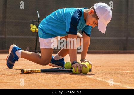Little boy in sportswear and cap playing tennis on a dirt tennis court. He picks up the tennis balls and puts them together on the racket Stock Photo