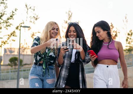 Three young female friends walking through the street using smartphone and having fun Stock Photo
