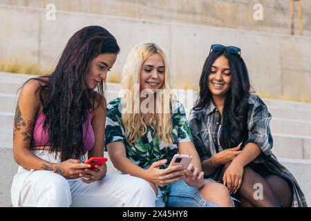 Three young multiethnic friends sitting on some steps outdoors using smartphone and having fun Stock Photo