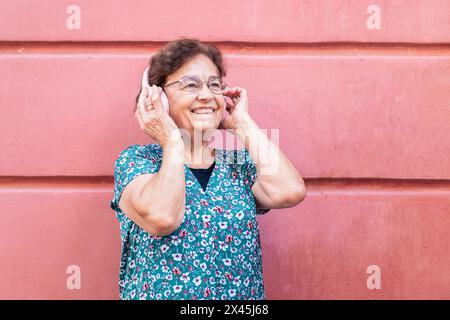 Smiling old lady in colorful clothes listening to music in modern pink headphones. Pink background Stock Photo