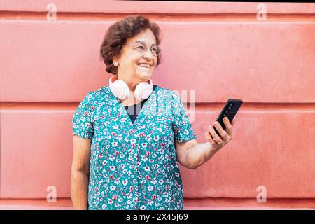 Smiling senior woman in colorful clothes using the smartphone with modern pink headphones in her neck. Pink background Stock Photo