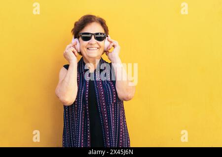 Smiling old woman in colorful clothes and sunglasses listening to music in modern pink headphones. Yellow background Stock Photo