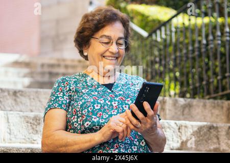 Smiling senior woman in colorful clothes walking down the street while using a smart phone Stock Photo