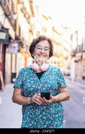 Portrait of a smiling senior woman in colorful clothes outdoors using a smartphone and having modern pink headphones attached to her neck Stock Photo