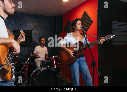musicians rehearsing their songs in a music rehearsal room. A girl sings and plays the guitar. One boy plays the drums and another one plays the bass Stock Photo