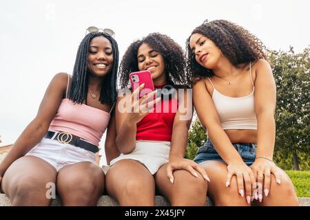 Three African American girls with afro hair sitting in a park laughing and having a good time and using smartphone Stock Photo