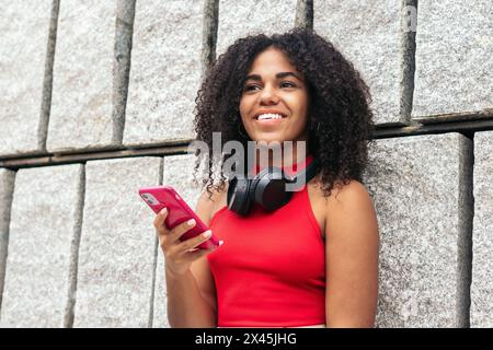 Portrait of a girl with afro hair in colorful summer clothes using the smartphone and wearing headphones on her neck Stock Photo
