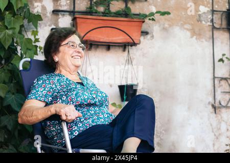 Calm and relaxed elderly woman sitting smiling on a chair in her backyard. She enjoys a humble summer Stock Photo