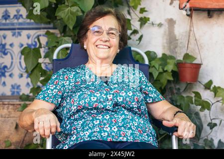 Calm and relaxed elderly woman sitting smiling on a chair in her backyard. She enjoys a humble summer Stock Photo