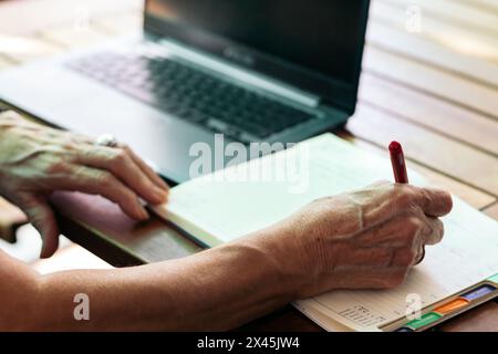 Close up of an unrecognizable old woman writing in a notebook while using her laptop at home. Stock Photo