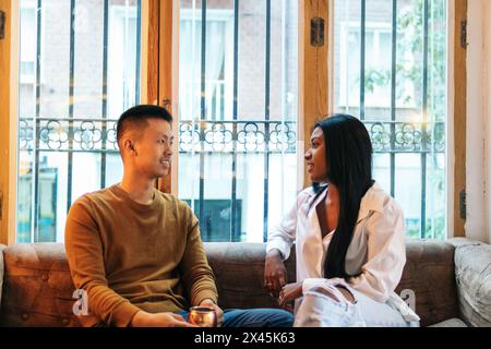 Asian man and Latin American woman sitting on the sofa in an interior space with warm light. They talk relaxed while having a drink. Stock Photo