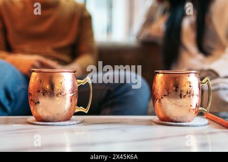 Close-up of two vintage-style copper mugs for the Moscow Mule. In the background of the image two people sitting on a sofa. Stock Photo