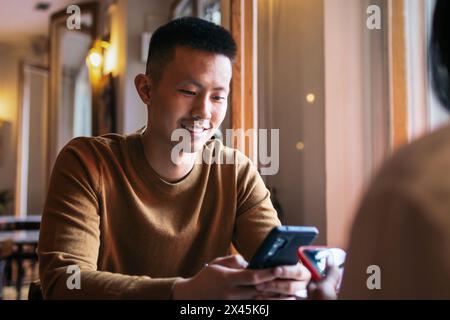 Asian man in casual clothes sitting in indoor cafe drinking coffee and using smartphone. Stock Photo