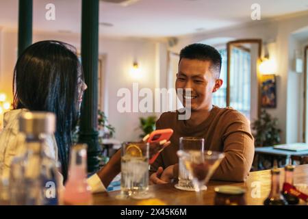 Young asian man and latin woman having a cocktail at a bar counter talking relaxed and having fun using smartphone. She shows him her social media on Stock Photo