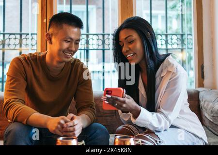Young asian man and latin woman sitting on sofa talking relaxed using smartphone. She shows him her social media on the screen Stock Photo