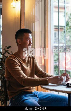 Asian man sitting at a coffee shop table drinking coffee. He smiles and has a good time talking with his companion Stock Photo