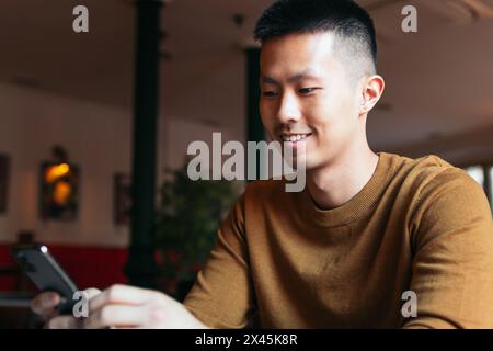Close up of smiling Asian man sitting in indoor coffee shop using smartphone. Stock Photo