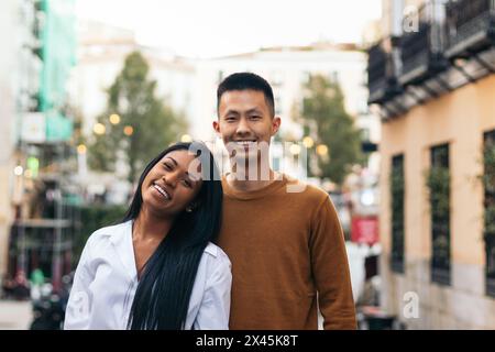 Interracial couple portrait: Asian man and Latin American woman embracing looking at camera smiling. They are standing on the city street wearing casu Stock Photo